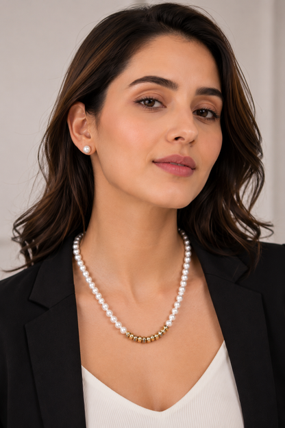 A woman wearing a double-strand white pearl necklace with a gold-plated connector link, styled with a small matching pearl stud earring and a black blazer.